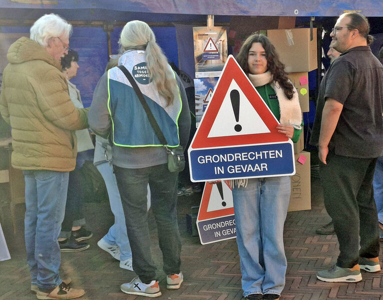 Groepje mensen, waarvan jonge vrouw een gevaarsbord met onderschrift 'Grondrechten in gevaar' vasthoudt.