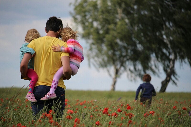 Vader met twee kinderen op armen wandelt door graanveld achter ouder kind