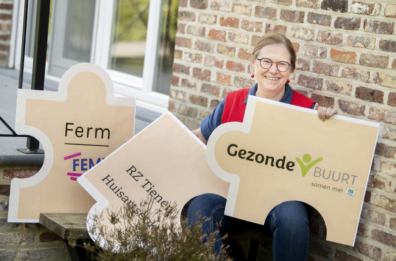 Annemie Steutelings houdt 3 grote puzzelstukken vast met logo's van Gezonde Buurt, Femma, Ferm en RZ Tienen.