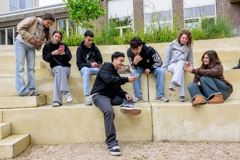Jongeren zitten samen buiten op een trapconstructie op school en zijn bezig met hun telefoon.